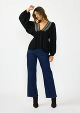 [Color: Black] A side facing image of a brunette model wearing a black long-sleeve blouse with a V-neckline and smocked trim, paired with a dark wash denim.