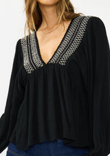 [Color: Black] A detail image of a brunette model wearing a black long-sleeve blouse with a V-neckline and smocked trim, paired with a dark wash denim.