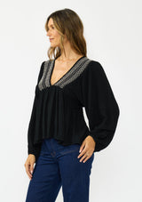 [Color: Black] A side facing image of a brunette model wearing a black long-sleeve blouse with a V-neckline and smocked trim, paired with a dark wash denim.