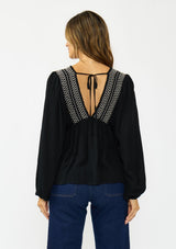 [Color: Black] A back facing image of a brunette model wearing a black long-sleeve blouse with a V-neckline, smocked trim, and a tie back detail, paired with a dark wash denim.