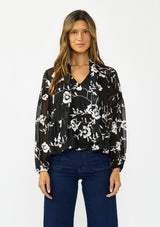 [Color: Black] A front facing image of a brunette model wearing a white and black blouse with floral embroidery and a tie neck detail. 