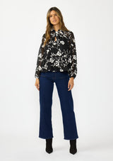 [Color: Black] A front facing image of a brunette model wearing a white and black blouse with floral embroidery and a tie neck detail. Styled with a dark wash denim and black boots. 