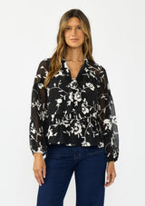 [Color: Black] A front facing image of a brunette model wearing a white and black blouse with floral embroidery and a tie neck detail. 