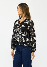 [Color: Black] A side facing image of a brunette model wearing a white and black blouse with floral embroidery and a tie neck detail. 