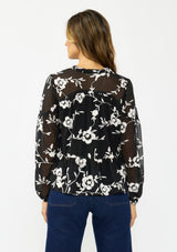 [Color: Black] A back facing image of a brunette model wearing a white and black blouse with floral embroidery and a tie neck detail. 