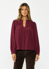 [Color: Merlot] A front facing image of a brunette model wearing a burgundy button up blouse with a v neckline and pintuck details. 