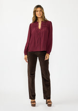 [Color: Merlot] A front facing image of a brunette model wearing a burgundy button up blouse with a v neckline and pintuck details. Styled with dark brown corduroys and brown platform heels. 