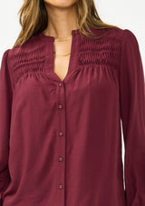 [Color: Merlot] A detail image of a brunette model wearing a burgundy button up blouse with a v neckline and pintuck details. 