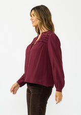 [Color: Merlot] A side facing image of a brunette model wearing a burgundy button up blouse with a v neckline and pintuck details. 