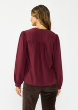 [Color: Merlot] A back facing image of a brunette model wearing a burgundy button up blouse with a v neckline and pintuck details. 