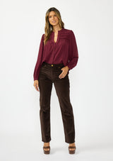 [Color: Merlot] A front facing image of a brunette model wearing a burgundy button up blouse with a v neckline and pintuck details. Styled with a dark brown corduroys and brown platform heels. 