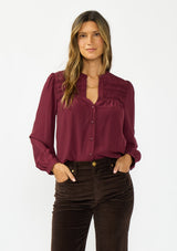 [Color: Merlot] A front facing image of a brunette model wearing a burgundy button up blouse with a v neckline and pintuck details. 
