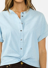 [Color: Light Denim Blue] A detail image of a brunette model wearing a light blue cotton top with short dolman sleeves, a button front, and a twist front hemline. Styled with brown corduroy bottoms. 