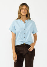 [Color: Light Denim Blue] A front facing image of a brunette model wearing a light blue cotton top with short dolman sleeves, a button front, and a twist front hemline. 