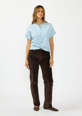[Color: Light Denim Blue] A front facing image of a brunette model wearing a light blue cotton top with short dolman sleeves, a button front, and a twist front hemline. Styled with brown corduroy bottoms. 