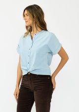 [Color: Light Denim Blue] A side facing image of a brunette model wearing a light blue cotton top with short dolman sleeves, a button front, and a twist front hemline. Styled with brown corduroy bottoms. 