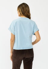 [Color: Light Denim Blue] A back facing image of a brunette model wearing a light blue cotton top with short dolman sleeves, a button front, and a twist front hemline. Styled with brown corduroy bottoms. 