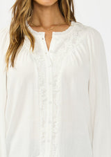 [Color: Petal] A detail image of a brunette model wearing a white blouse with embroidered lace, a button front, and long flowy sleeves. 