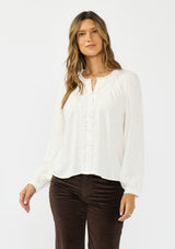[Color: Petal] A front facing image of a brunette model wearing a white blouse with embroidered lace, a button front, and long flowy sleeves. Styled with dark brown corduroy pants. 