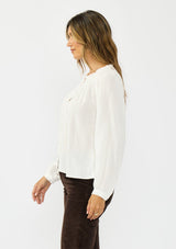 [Color: Petal] A front facing image of a brunette model wearing a white blouse with embroidered lace, a button front, and long flowy sleeves. Styled with dark brown corduroy pants. 