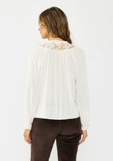 [Color: Petal] A back facing image of a brunette model wearing a white blouse with embroidered lace, a button front, and long flowy sleeves. Styled with dark brown corduroy pants. 
