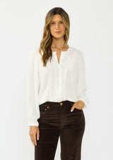 [Color: Petal] A front facing image of a brunette model wearing a white blouse with embroidered lace, a button front, and long flowy sleeves. Styled with dark brown corduroy pants. 