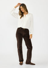[Color: Petal] A front facing image of a brunette model wearing a white blouse with embroidered lace, a button front, and long flowy sleeves. Styled with dark brown corduroy pants. 