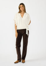 [Color: Vanilla] A front facing image of a brunette model wearing an ivory embroidered surplice top with flutter sleeves, an elastic hemline, and a tie detail. Styled with dark brown corduroys. 