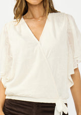 [Color: Vanilla] A detail image of a brunette model wearing an ivory embroidered surplice top with flutter sleeves, an elastic hemline, and a tie detail. Styled with dark brown corduroys. 