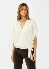 [Color: Vanilla] A front facing image of a brunette model wearing an ivory embroidered surplice top with flutter sleeves, an elastic hemline, and a tie detail. Styled with dark brown corduroys. 