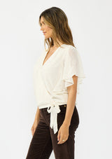 [Color: Vanilla] A side facing image of a brunette model wearing an ivory embroidered surplice top with flutter sleeves, an elastic hemline, and a tie detail. Styled with dark brown corduroys. 