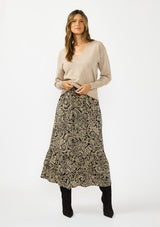 [Color: Black/Natural] A woman modeling a beige long-sleeve V-neck sweater paired with a black and beige paisley patterned midi skirt and black boots.