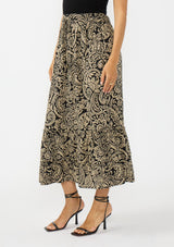 [Color: Black/Natural] A side facing image of a model wearing a tan and lack boho paisley skirt with a tiered ruffle hem. Styled with black strappy heels. 
