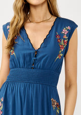 [Color: Navy] A detail image of a blonde model wearing a blue embroidered maxi dress with w scalloped v-neckline, a smocked waist, and slits. A flowy summer dress for garden parties and soirees. 