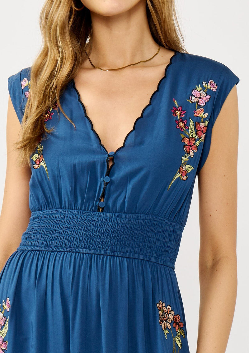 [Color: Navy] A detail image of a blonde model wearing a blue embroidered maxi dress with w scalloped v-neckline, a smocked waist, and slits. A flowy summer dress for garden parties and soirees. 