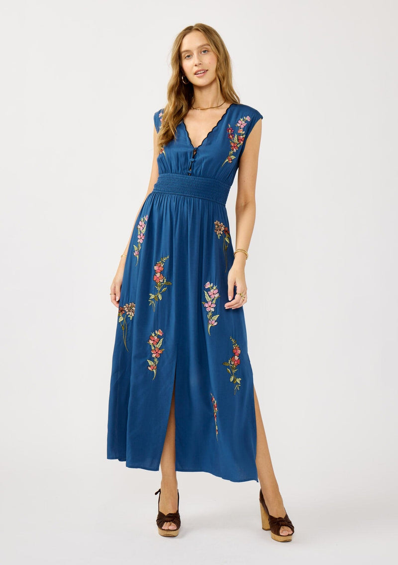 [Color: Navy] A front facing image of a blonde model wearing a blue embroidered maxi dress with w scalloped v-neckline, a smocked waist, and slits. A flowy summer dress for garden parties and soirees. 