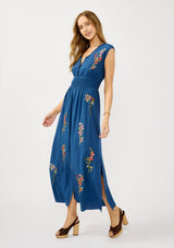 [Color: Navy] A side facing image of a blonde model wearing a blue embroidered maxi dress with w scalloped v-neckline, a smocked waist, and slits. A flowy summer dress for garden parties and soirees. 