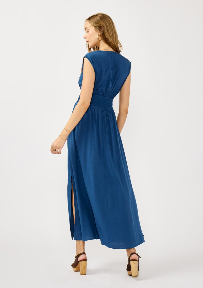[Color: Navy] A back facing image of a blonde model wearing a blue embroidered maxi dress with w scalloped v-neckline, a smocked waist, and slits. A flowy summer dress for garden parties and soirees. 