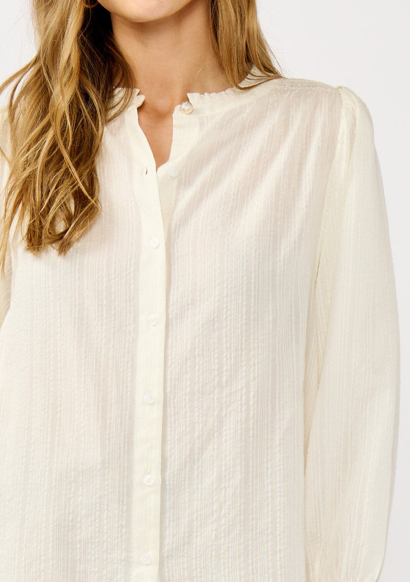 [Color: Cream] A detail image of a blonde model wearing an ivory button front top with a high ruffled neckline, long sleeves, and smocked elastic cuffs. A relaxed fit blouse perfect for the office and special occasions. 