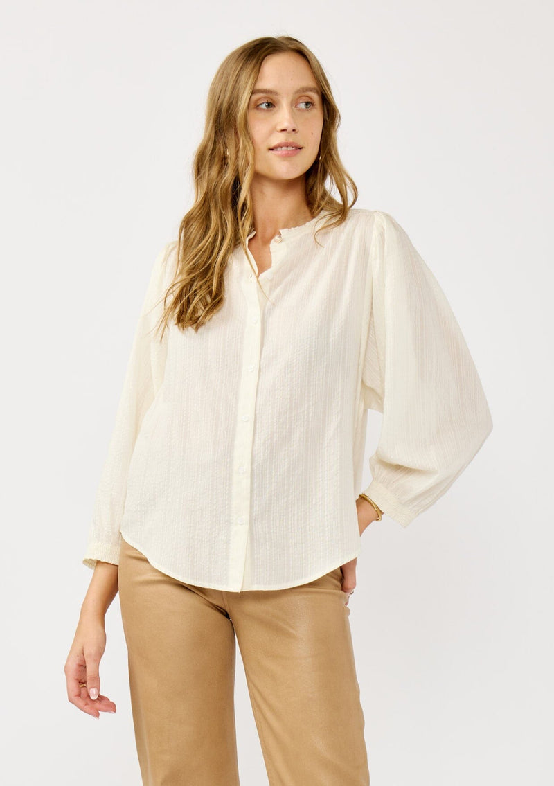 [Color: Cream] A front facing image of a blonde model wearing an ivory button front top with a high ruffled neckline, long sleeves, and smocked elastic cuffs. A relaxed fit blouse perfect for the office and special occasions. 