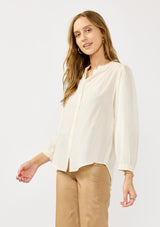 [Color: Cream] A side facing image of a blonde model wearing an ivory button front top with a high ruffled neckline, long sleeves, and smocked elastic cuffs. A relaxed fit blouse perfect for the office and special occasions. 