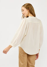 [Color: Cream] A back facing image of a blonde model wearing an ivory button front top with a high ruffled neckline, long sleeves, and smocked elastic cuffs. A relaxed fit blouse perfect for the office and special occasions. 
