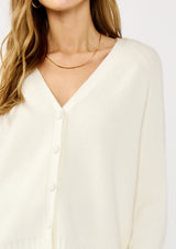 [Color: White] A detail image of a blonde model wearing a white cozy rib stitch cardigan with a button front, v-neckline, ribbed border, and cable knit detailing along the sleeves. 