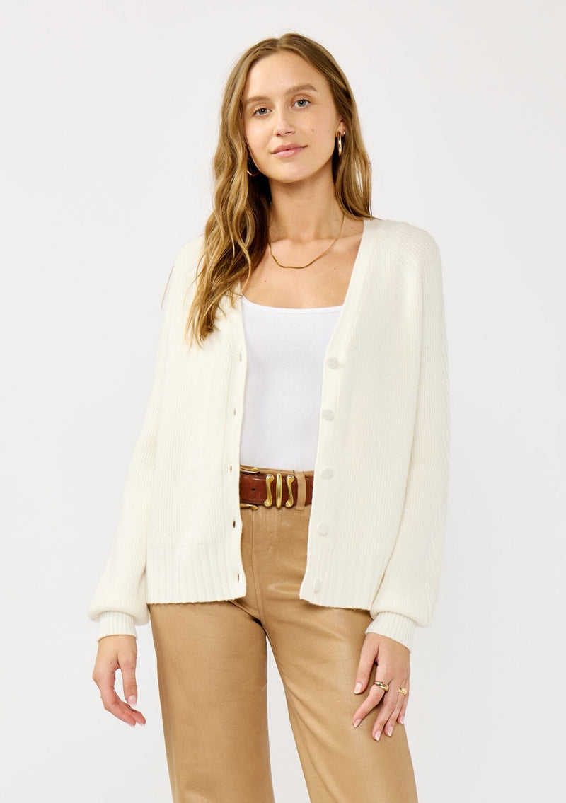 [Color: White] A front facing image of a blonde model wearing a white cozy rib stitch cardigan with a button front, v-neckline, ribbed border, and cable knit detailing along the sleeves. 
