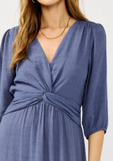 [Color: Slate Blue] A detail image of a blonde model wearing a blue flowy maxi dress with long sleeves, a twist front, v-neckline, and a high low hemline. A special occasion dress for day and night. 