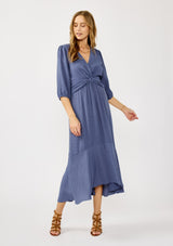 [Color: Slate Blue] A front facing image of a blonde model wearing a blue flowy maxi dress with long sleeves, a twist front, v-neckline, and a high low hemline. A special occasion dress for day and night. 