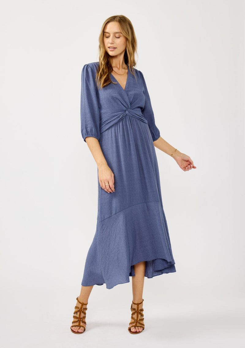 [Color: Slate Blue] A front facing image of a blonde model wearing a blue flowy maxi dress with long sleeves, a twist front, v-neckline, and a high low hemline. A special occasion dress for day and night. 