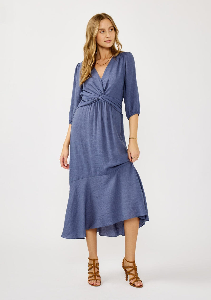 [Color: Slate Blue] A front facing image of a blonde model wearing a blue flowy maxi dress with long sleeves, a twist front, v-neckline, and a high low hemline. A special occasion dress for day and night. 