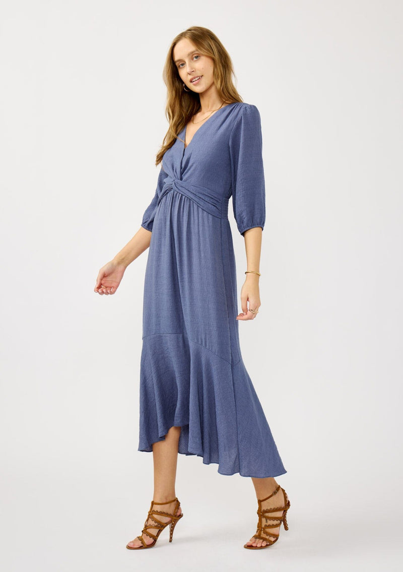 [Color: Slate Blue] A side facing image of a blonde model wearing a blue flowy maxi dress with long sleeves, a twist front, v-neckline, and a high low hemline. A special occasion dress for day and night. 