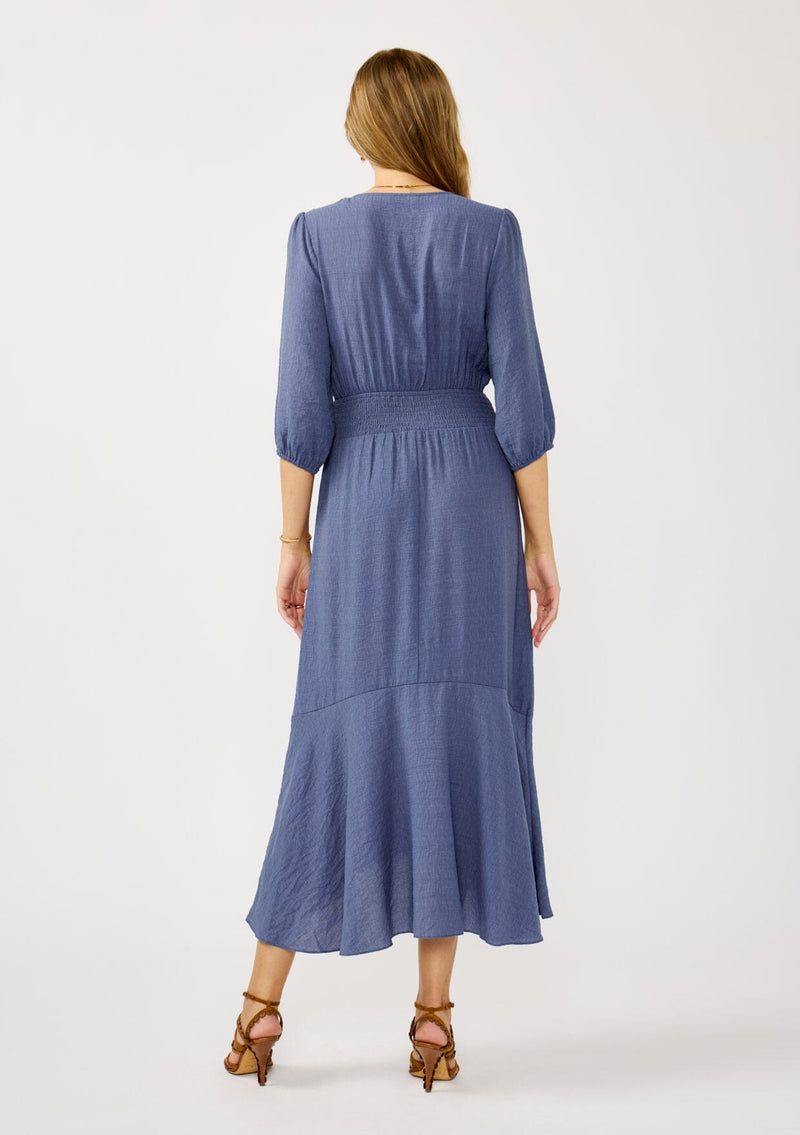 [Color: Slate Blue] A back facing image of a blonde model wearing a blue flowy maxi dress with long sleeves, a twist front, v-neckline, smocked back, and a high low hemline. A special occasion dress for day and night. 